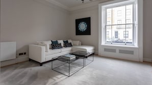 A spacious living room with a white sofa, a glass coffee table, and a large window with a view of buildings.