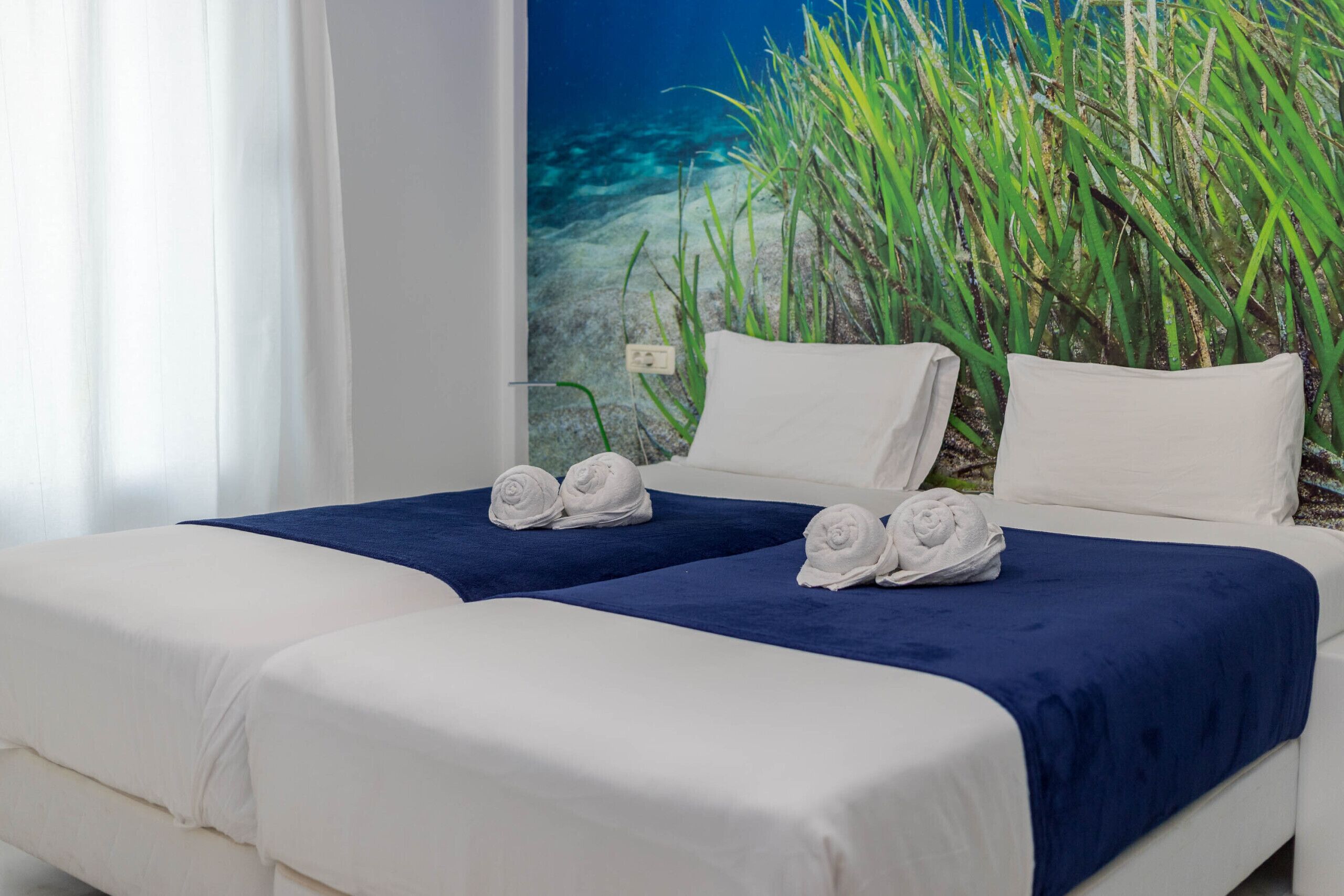 A hotel room with two beds, each with white linens and a blue accent. There are towels on the beds and a mural of underwater plants on the wall.