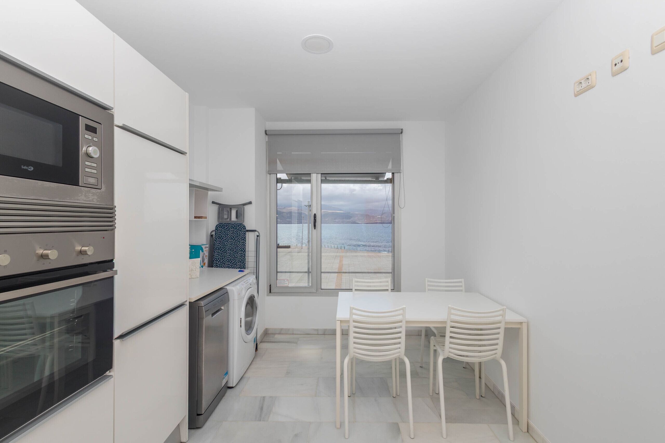 Une cuisine moderne équipée d’un four, d’un micro-ondes et d’un lave-linge intégrés. Elle comprend un coin repas avec une table et des chaises, et offre une vue sur la mer grâce à une grande fenêtre.