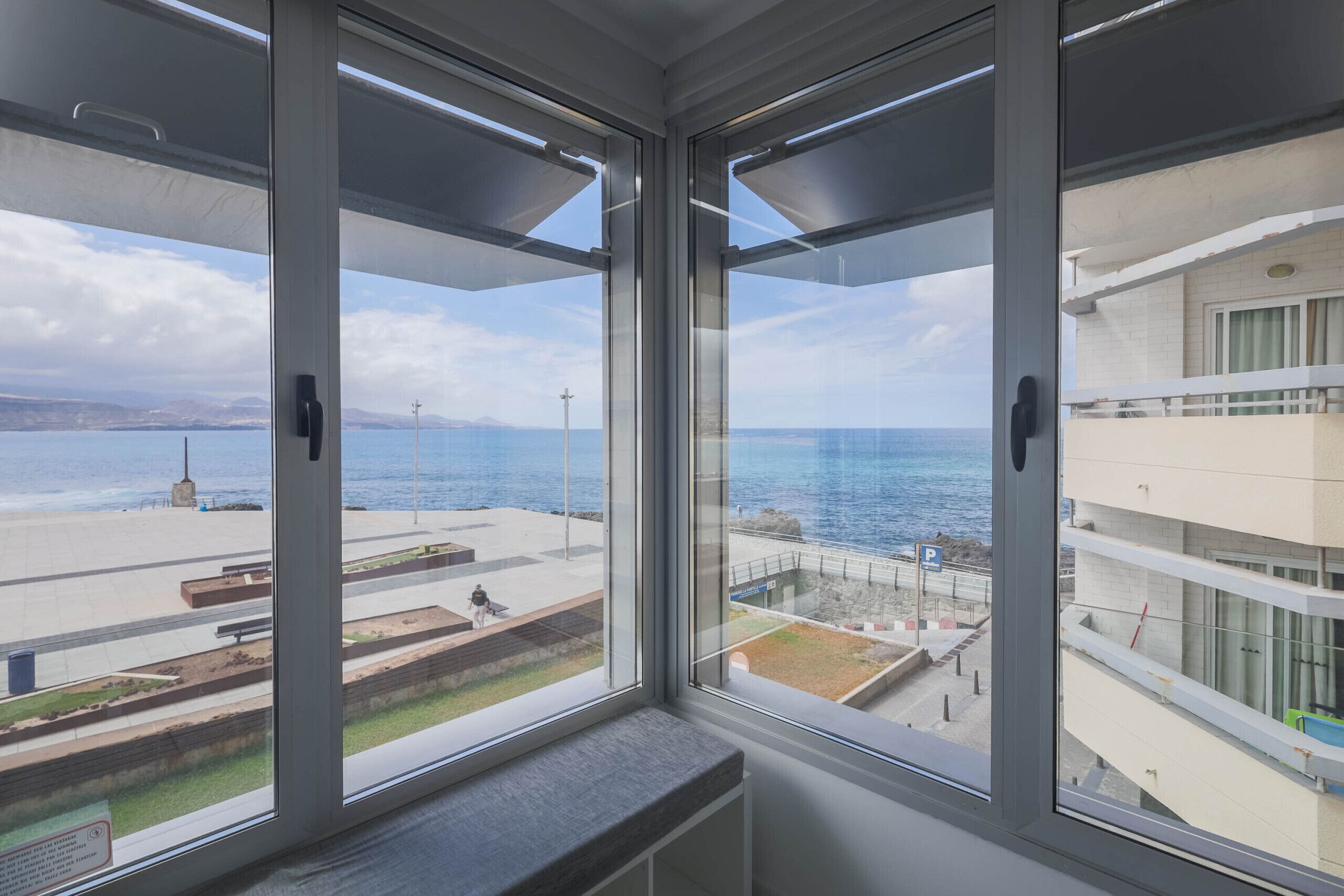 A room with a large window overlooking a coastal promenade with a view of the sea and a building across the promenade.