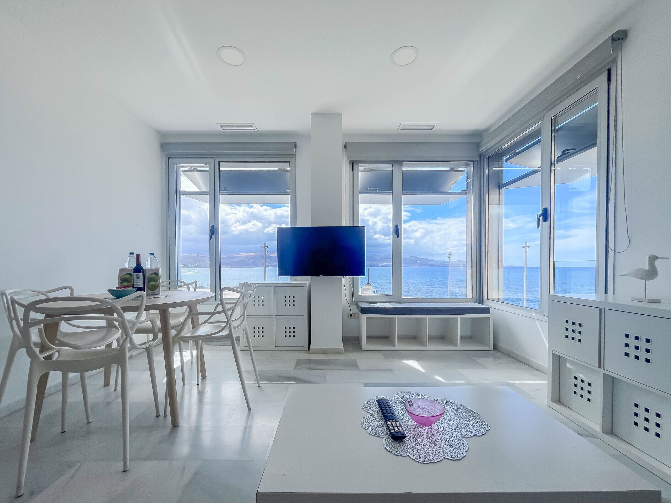 A modern living room with a large window, a flat-screen TV, white furniture, and a view of the sea.