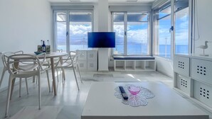 Moderni olohuone, jossa on suuri ikkuna, litteänäyttöinen televisio, valkoiset huonekalut ja näkymä merelle.