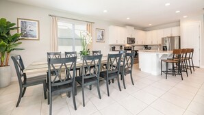 A modern kitchen with white cabinets, a central island with bar stools, and a dining area with a glass table and dark chairs. - 25% Off Encore Resort Arcade Home (Kissimmee)