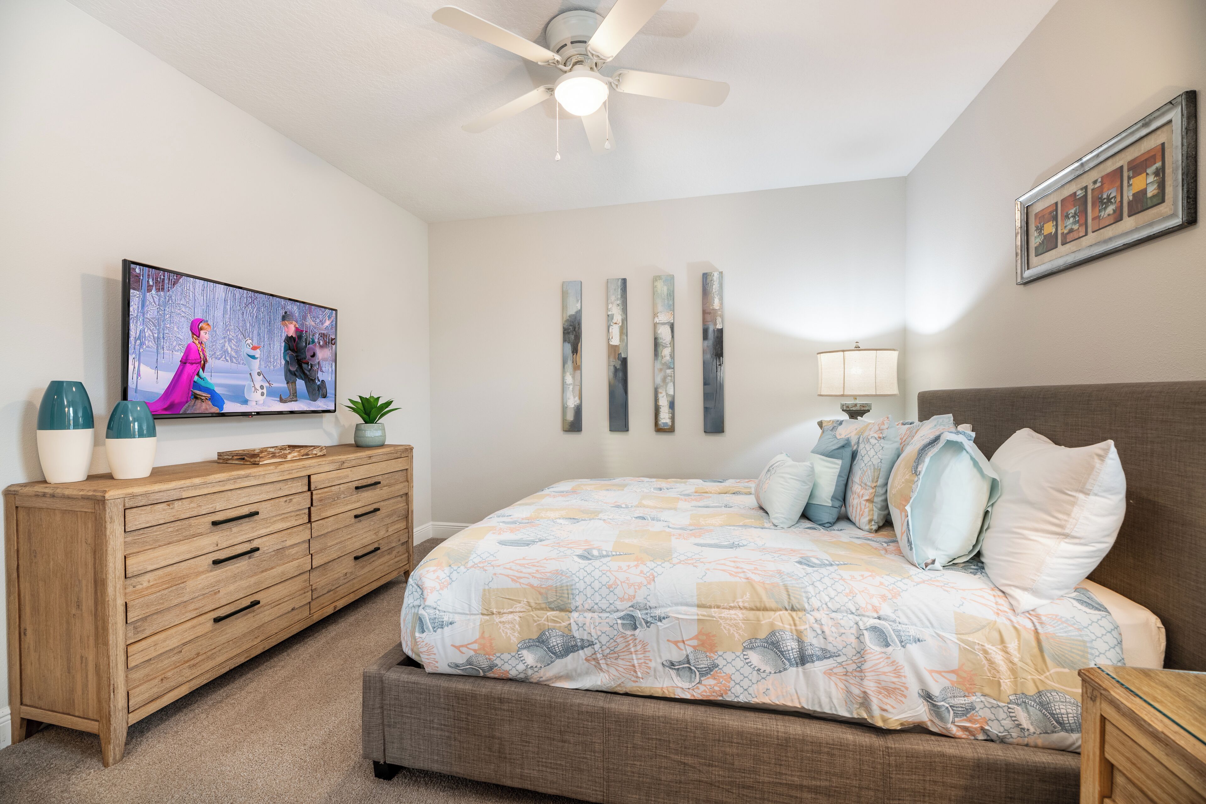 A bedroom with a bed, a TV, a ceiling fan, a dresser, and artwork on the wall.