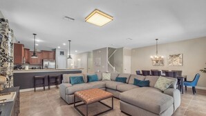 A spacious living room with a large sectional sofa, a coffee table, and a kitchen area with wooden cabinets and a refrigerator.