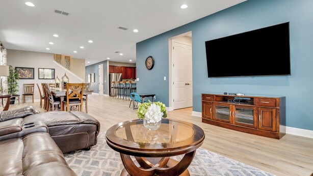 A modern living room with a sectional sofa, a glass-top coffee table, a flat-screen TV mounted on the wall, and a dining area in the background. - 25% Off 10BR Encore Pool Villa Near DIsney (Kissimmee)