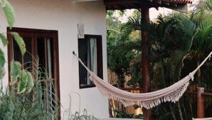 Una hamaca atada entre postes de madera en un jardín con terraza de madera, rodeada de exuberante vegetación y un edificio con ventana y puerta.