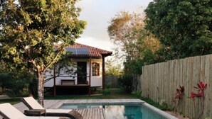 Un patio trasero con piscina, terraza de madera, sillas de descanso y una casa pequeña, todo rodeado por un cerco de madera.