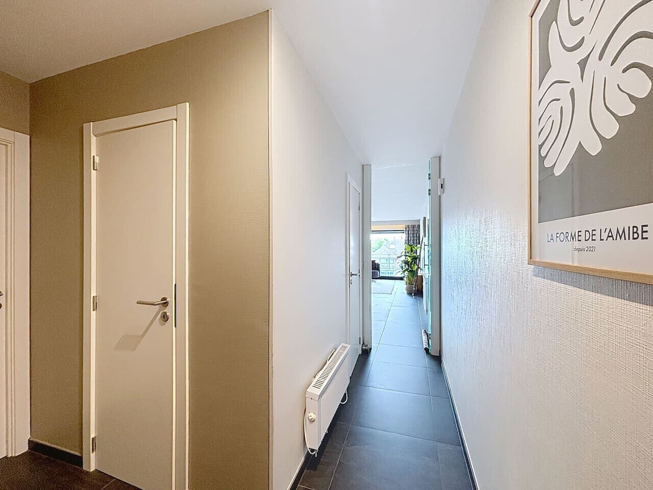 A hallway with a door on the left, a radiator on the right, and a framed artwork on the wall.