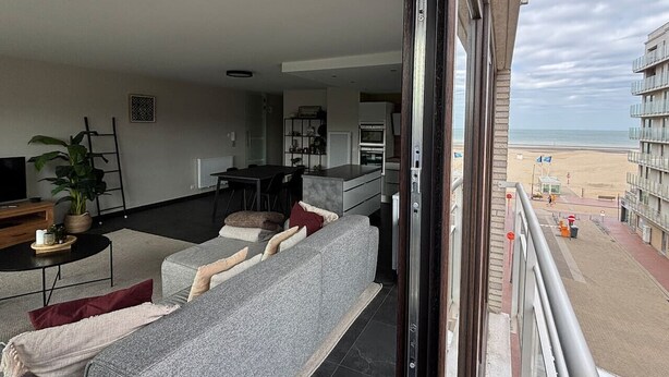 A modern living room with a grey sofa, a coffee table, and a TV. A balcony overlooks a beach with a building in the background. - Flat Zandloper - Side sea view in Oostduinkerke (Koksijde)