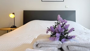 A neatly made bed with folded white towels and a purple flower arrangement on the bed. - Flat Zandloper - Side sea view in Oostduinkerke (Koksijde)