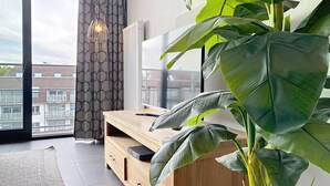 A modern living room with a large plant, a wooden TV stand, and a view of the city through a large window. - Flat Zandloper - Side sea view in Oostduinkerke (Koksijde)