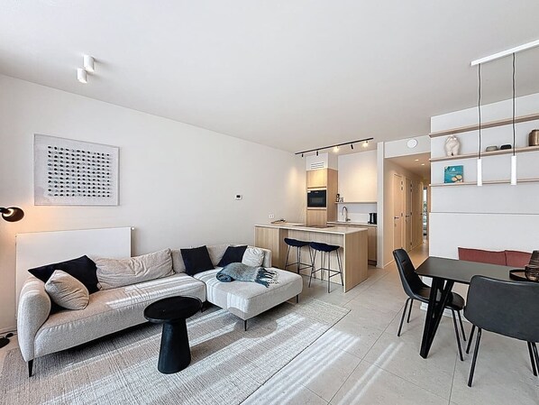 A modern living room with a sofa, ottoman, and coffee table, a kitchen area with a dining table and chairs, and a small bar area.