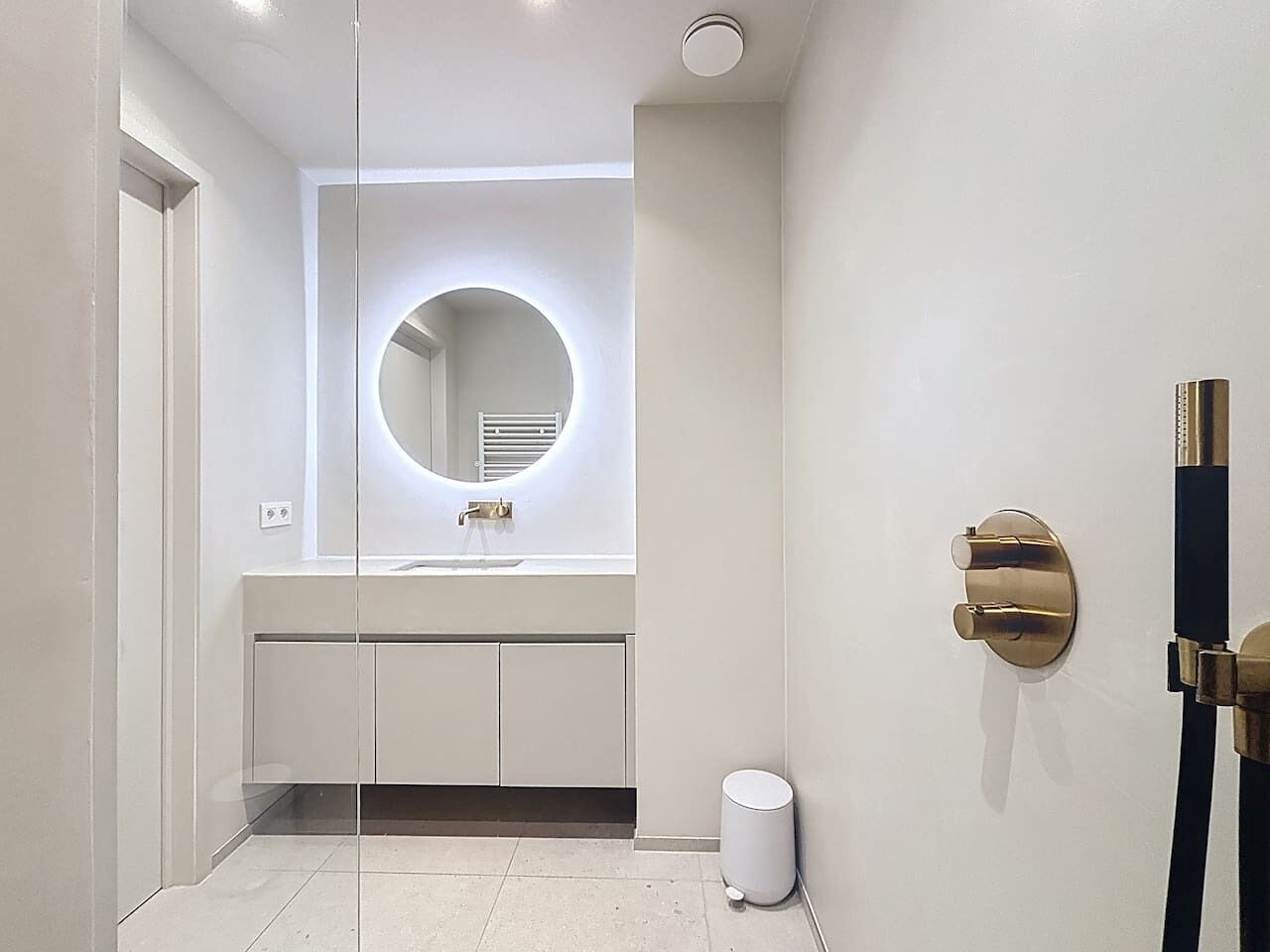 Een moderne badkamer met een glazen wand, een ronde spiegel, een witte wastafel en een douche met een gouden kraan.