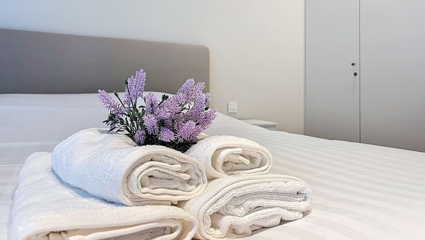 A neatly made bed with rolled white towels and a small purple flower arrangement.