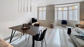 A modern living room with a dining area, large windows with sheer curtains, a flat-screen TV, and a blue chair.