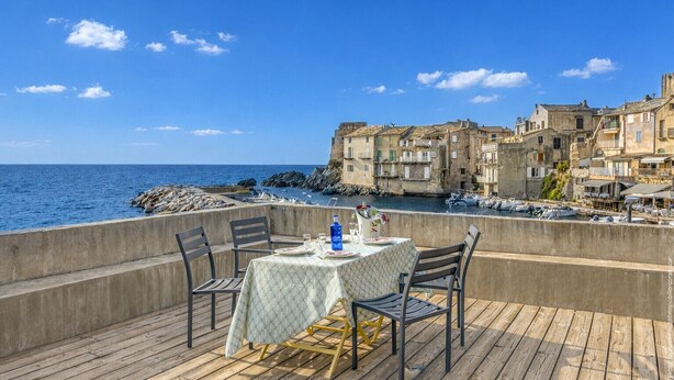 A terrace with a table set for two, overlooking a coastal town with historic buildings.