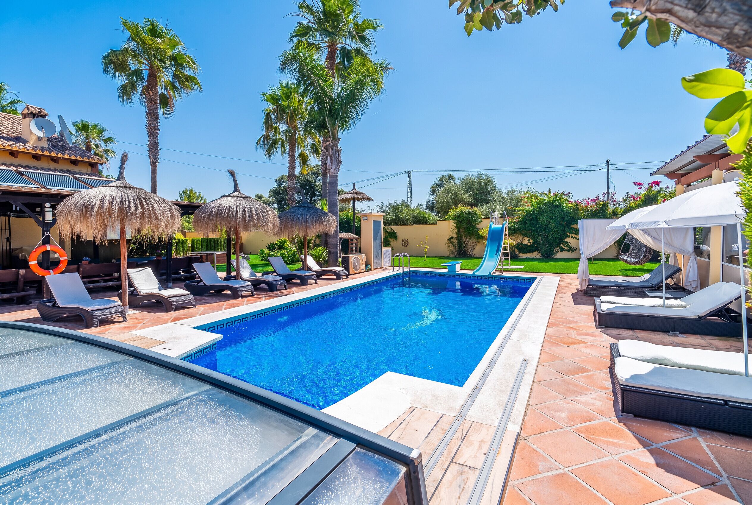 A swimming pool with lounge chairs and umbrellas, surrounded by palm trees and a clear sky.