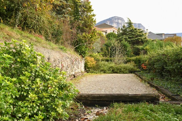 A gravel path leading to a wooden bridge over a stream, surrounded by lush greenery and a mountain in the background.