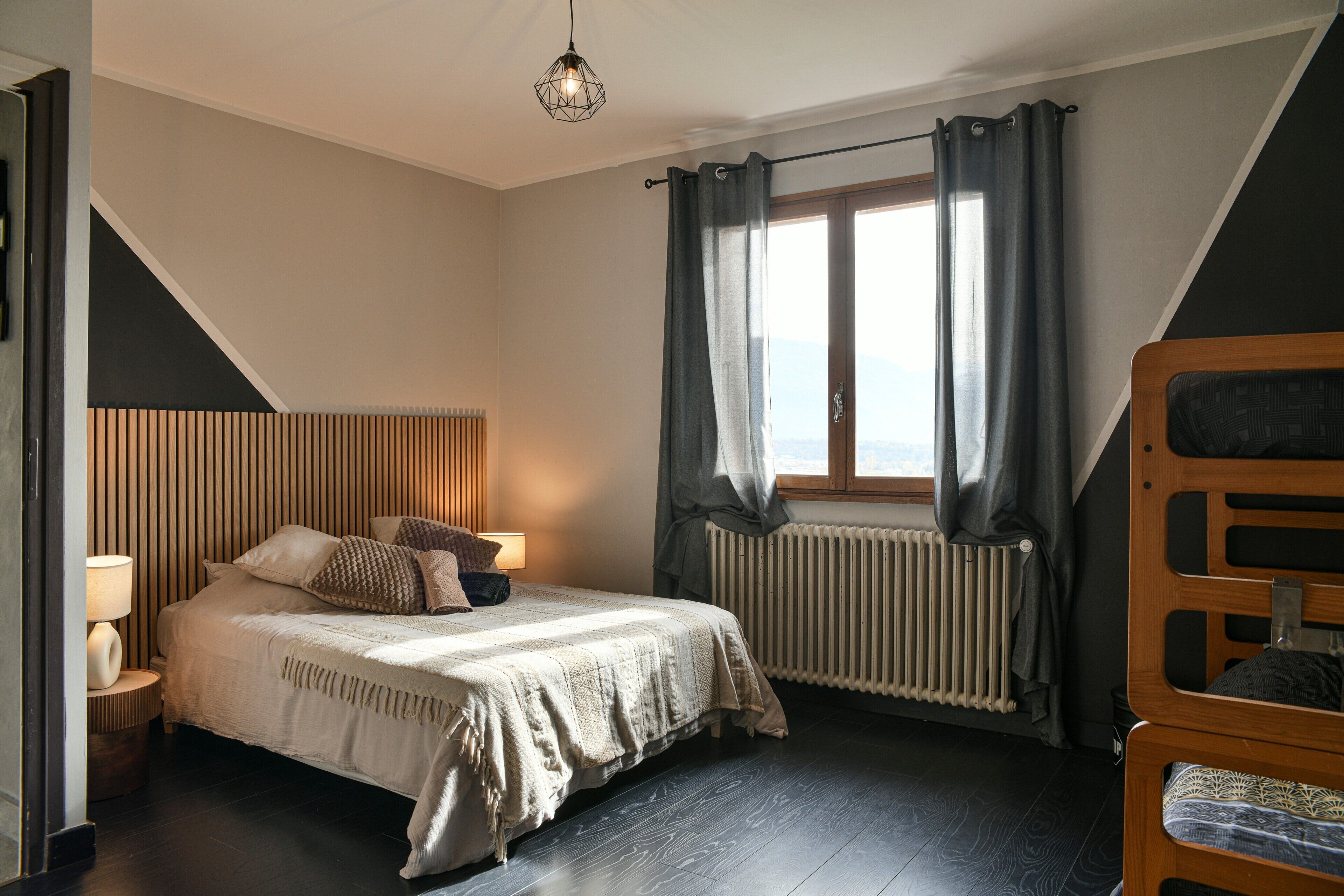 A bedroom with a bed, a bedside lamp, a window with curtains, a radiator, and a bunk bed.