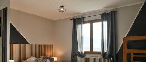 A bedroom with a bed, a bedside lamp, a window with curtains, a radiator, and a bunk bed.