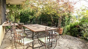 Un patio al aire libre con una mesa de madera y sillas de metal, rodeado de abundante vegetación y un piso de piedra.