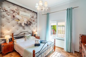 A bedroom with a tufted headboard, a chandelier, a bed with white linens, a nightstand with a lamp, and a view of greenery through a window.