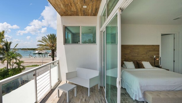 A balcony with a white sofa and a small table, a bed with white bedding, and a view of the sea and boats. - Miami Stunning Waterfront Villa Escape (North Bay Village)