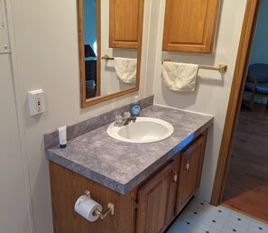 A bathroom with a marble countertop, a white sink, wooden cabinets, a mirror, and a towel rack. - Large 1-Bdrm+ Apartment Surrounded by Nature in Rural Vermont (Addison)
