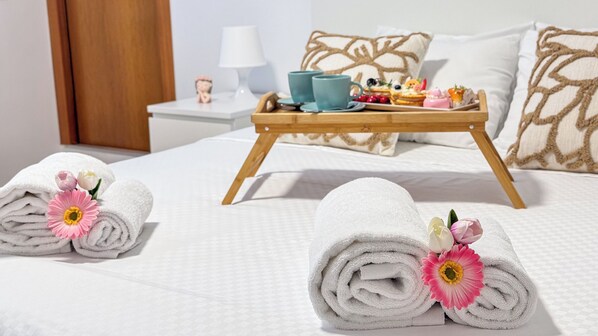 Una cama cuidadosamente tendida con sábanas blancas y toallas enrolladas decoradas con flores, una bandeja con tetera y pasteles, y una mesita de noche con lámpara y una figurita.