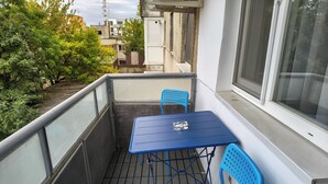 A balcony with a blue table and two blue chairs, a window, and a view of buildings and trees. - Student Grey Apartment w/ Full ammenities. Spacious 2 room Balcony (Timisoara)