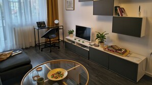 A modern living room with a sofa, a glass coffee table, a TV, a desk with a computer, and a bookshelf. - Student Grey Apartment w/ Full ammenities. Spacious 2 room Balcony (Timisoara)