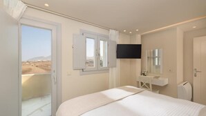 Ein Hotelzimmer mit Bett, Wandfernseher, einem Fenster mit Vorhängen, einem kleinen Schreibtisch mit Spiegel und Blick auf eine Wüstenlandschaft.
