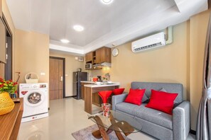 Un espacio habitable compacto con kitchenette, lavadora, sofá gris con cojines rojos y una mesa de centro de vidrio.
