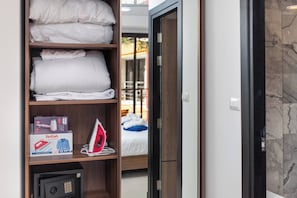 Habitación de hotel con sábanas blancas ordenadas en estantes, una cama a la vista con un cojín azul y un vaporizador Tefal en un estante.