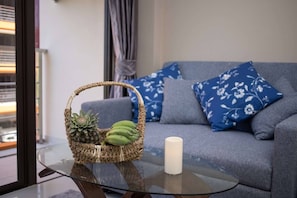 A room with a glass table, a wicker basket with a pineapple and bananas, a white candle, and a grey sofa with blue floral cushions.