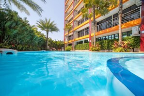 A swimming pool with clear blue water, surrounded by lush greenery and a multi-story building with balconies. - Nara Condo 1br | Pool view | nbc nai harn (Rawai)