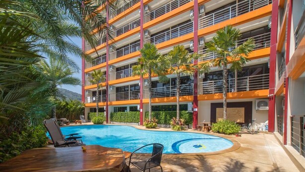 A multi-story hotel with a swimming pool, outdoor seating, and palm trees. - Nara Condo 1br | Pool view | nbc nai harn (Rawai)