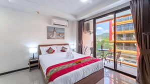 A hotel room with a large bed, a balcony with a table and chairs, and a view of a building with orange and white colors. - Nara Condo 1br | Pool view | nbc nai harn (Rawai)