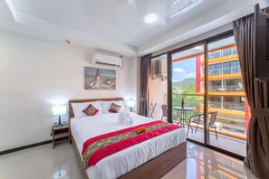 A hotel room with a large bed, a balcony with a table and chairs, and a view of a building with orange and white colors. - Nara Condo 1br | Pool view | nbc nai harn (Rawai)