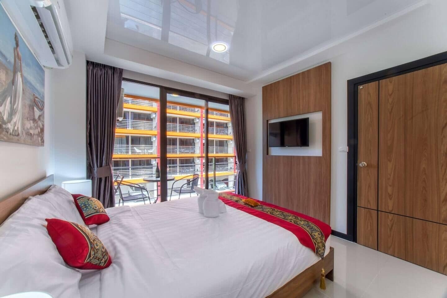 A hotel room with a bed, a TV mounted on the wall, and a view of an outdoor balcony with yellow railings.