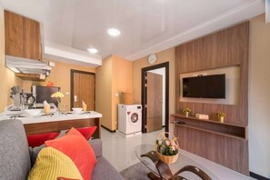 A modern living room with a sofa, a coffee table, a TV mounted on the wall, a washing machine, and a kitchen area with a sink and utensils.
