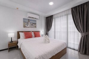 A hotel room with a bed, a nightstand, a lamp, an air conditioning unit, a painting, and large curtains.