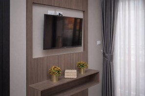 A flat-screen TV mounted on a wall with a wooden panel design, two small potted plants on a shelf below, and a window with sheer curtains. - Vero Condo 1br | Garden view | nbc nai harn (Rawai)