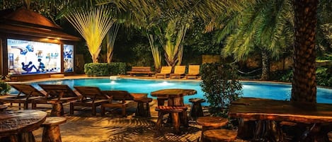 A poolside area with lounge chairs, wooden tables, and a bar area with illuminated artwork.