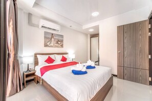 A hotel room with a large bed, a nightstand, a lamp, a painting on the wall, an air conditioning unit, and a wardrobe.