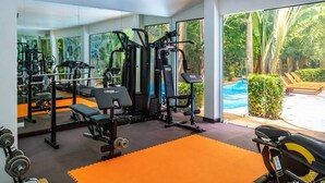 Et fitnessrum med forskellig træningsudstyr, en bænk og en swimmingpool, der ses gennem vinduerne.