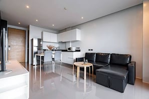 A modern living room with a black leather sofa, a dining area with a white table and chairs, and a kitchen with white cabinets and stainless steel appliances.