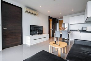 A modern living room with a flat-screen TV, a dining area with a round table, and a kitchen with white cabinets.