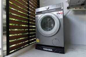 A Toshiba washing machine in a room with a window featuring horizontal blinds.